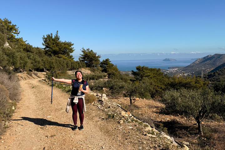 A landscape photo with Brenda in the bottom left corner red in the face with a hiking stick and a sandwich overlooking a beautiful vista of Crete's mountain and ocean beyond.