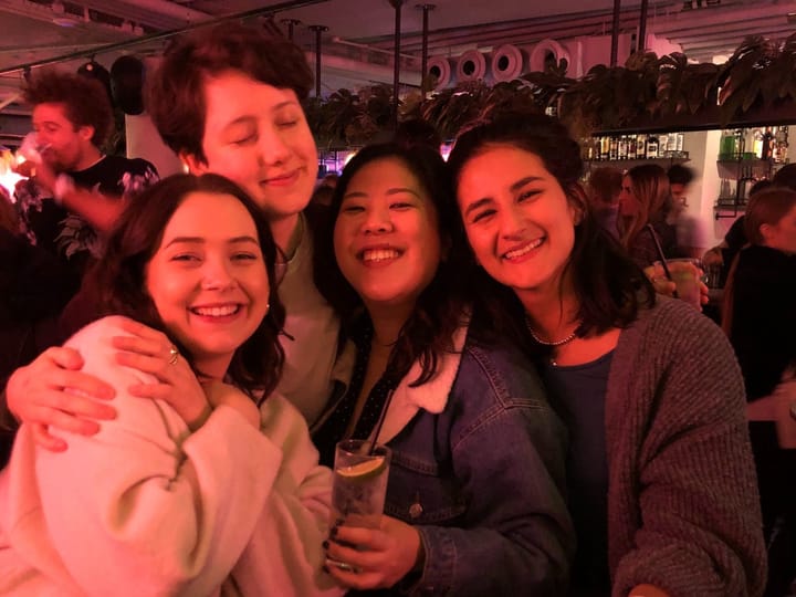 Four women at a bar. It's dimly lit with dusky red. Three of them are looking at the camera and smiling, and one of them has 