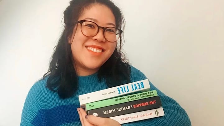 A very happy Brenda wearing glasses smiles wearing a turquoise jumper, looking into the camera. In her arm she's carrying a s