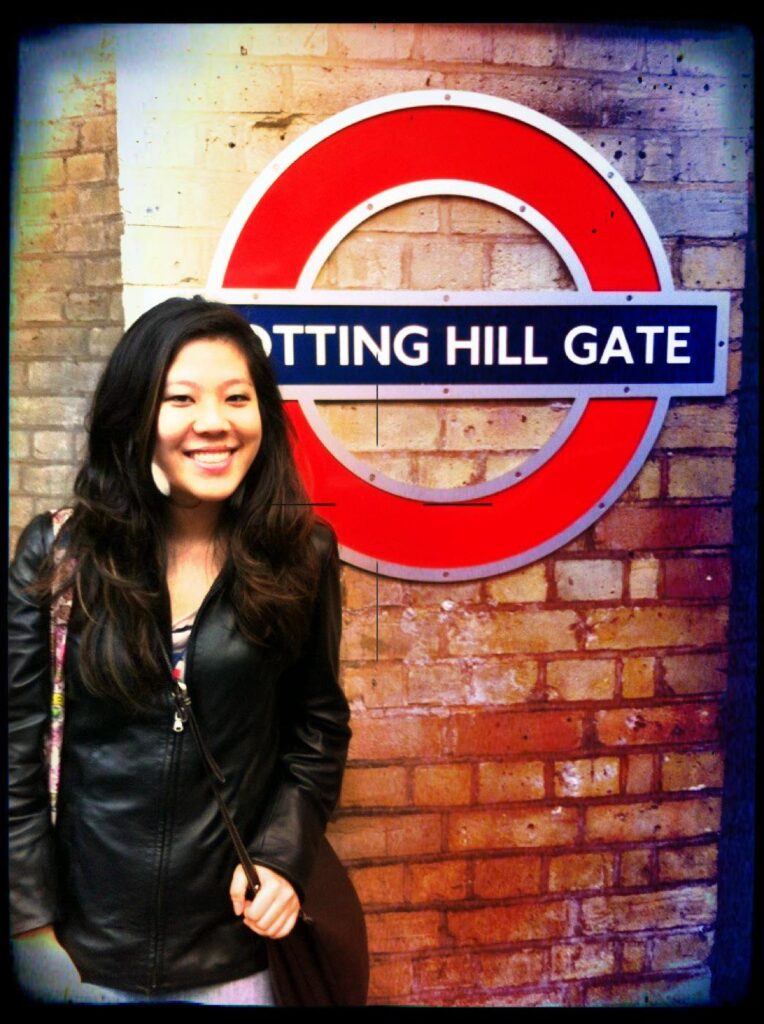 A photo of Brenda nine years ago on her first trip to London. She's smiling at the camera in front of a brick wall, the iconic Notting Hill Gate TFL Underground roundel behind her.