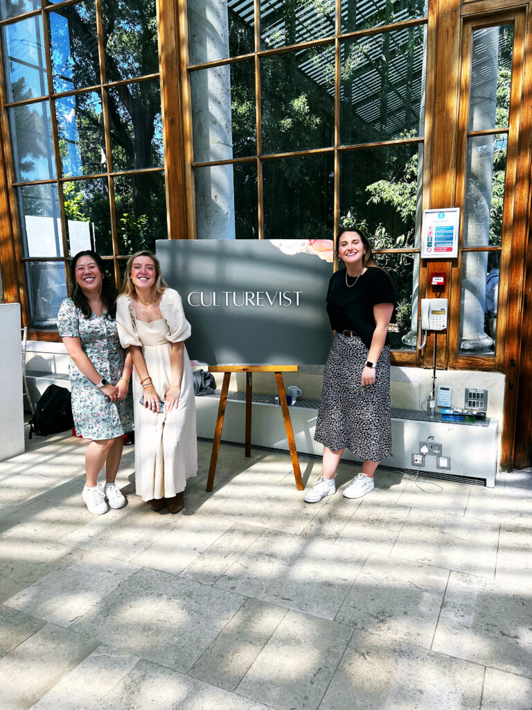 A photo of three women (left to right: Brenda, Charlotte, and Amy) smiling in front of a sign that says 'Culturevist'. This is them at the Culturevist Conference in 2022 at Kew Gardens, and they are smiling widely at the camera. 