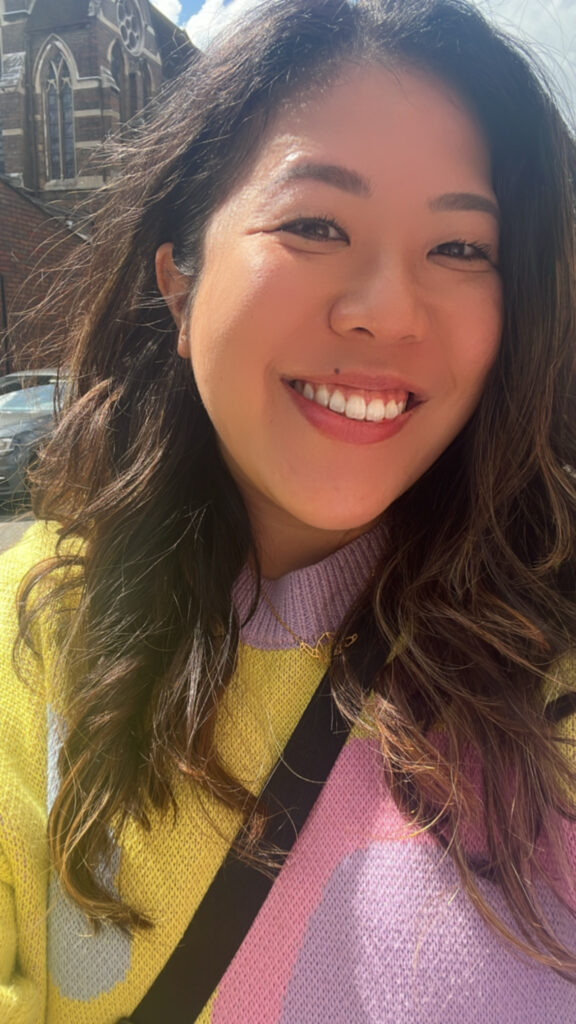 A selfie, a woman with long dark hair wearing a purple and yellow watercolour-syle jumper is smiling into the camera.