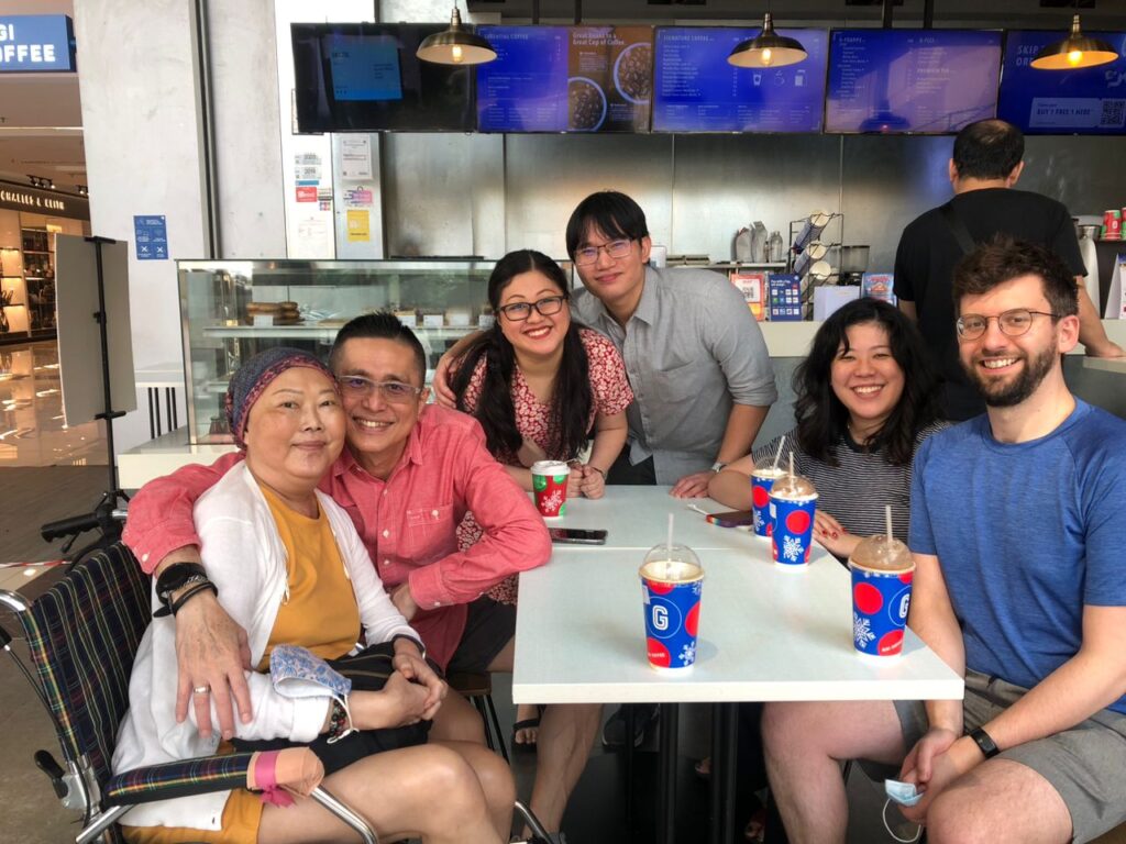 A photo of a family at a coffee shop, with various lattes on the table, all smiling at the camera. Mum is on the left in her wheelchair while her family surrounds her. 