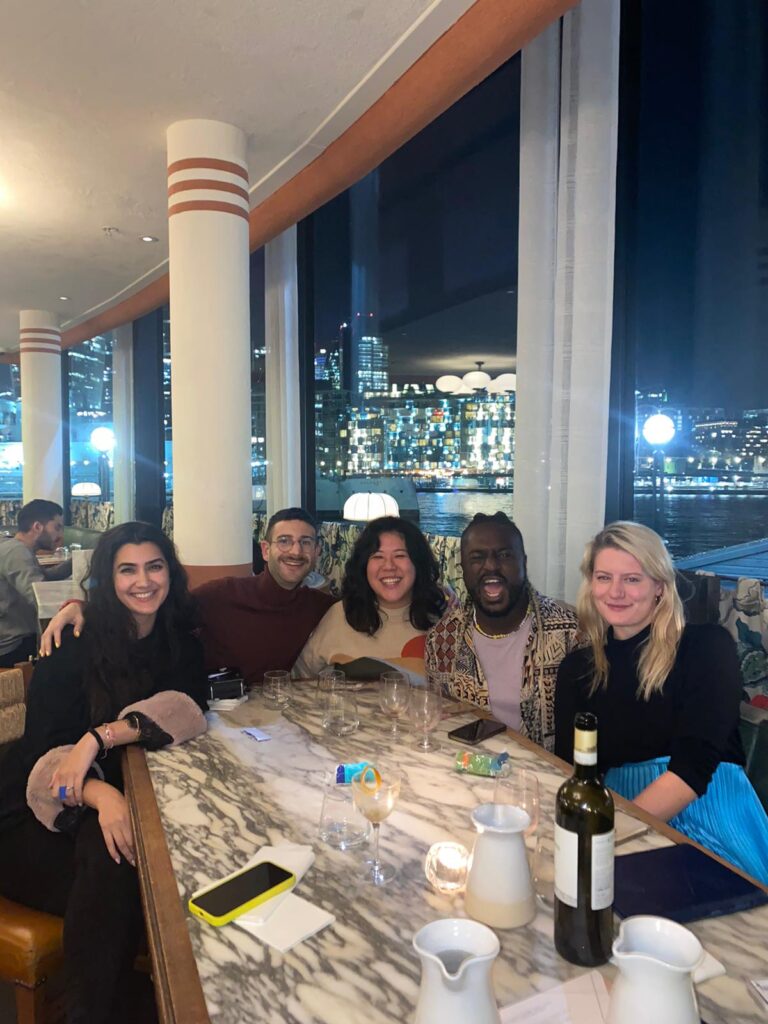 A group of five friends sit at a marbled dining table smiling into the camera. The backdrop is nighttime on the river, city lights making the water twinkle.